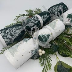 three rolled up christmas crackers sitting on top of green leaves and pine cones, tied with white ribbon