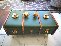 a green trunk with gold trimmings sitting on top of a tile floor next to two cups and saucers