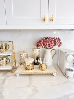 a white kitchen counter with gold accents and pink flowers in a vase on the shelf