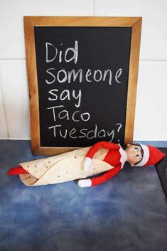 an elf laying on top of a table next to a chalkboard with the words did someone say taco tuesday?