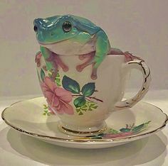 a green frog sitting on top of a white cup and saucer next to a plate