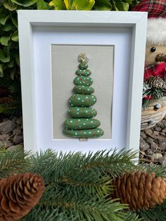 a green christmas tree in a white frame next to pine cones and evergreen branches with a teddy bear