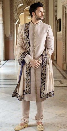 a man standing in a hallway wearing a beige outfit with blue accents and gold details