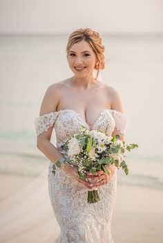 a woman in a wedding dress standing on the beach holding a bridal bouquet and smiling at the camera