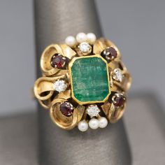 This knockout of a ring bring back visions of Renaissance jewelry- rich in colors and intricate in design. The ring features a center emerald cut emerald which is bezel set. The emerald has a closed back so we can't calculate the weight precisely, however it measures 10mm x 8.5mm, roughly calculating to 3.00ct. The stone is framed by four diamonds that measures 2.5mm each, totaling 0.24cttw. 2.8mm garnets accent the corners and the center top and bottom are highlighted by three seed pearls each. Pretty Jewelry, Seed Pearl, Bring Back, Pretty Jewellery, Bezel Setting, Emerald Cut, In Design, Statement Ring, Statement Rings