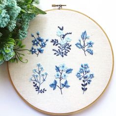 embroidered blue flowers are displayed on a white embroidery hoop next to a potted plant