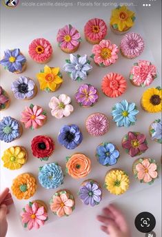 cupcakes decorated with fondant flowers are displayed on a white surface, and someone is placing them in the middle