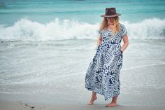 a woman in a dress and hat standing on the beach next to the ocean waves