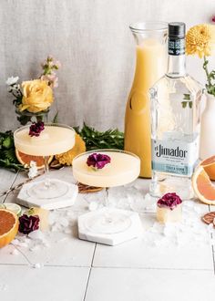 two glasses filled with drinks and garnished with flowers next to bottles of liquor