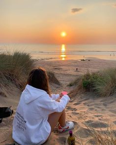 a woman sitting on top of a sandy beach next to the ocean at sunset,