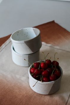 two white bowls filled with cherries on top of a cloth covered tablecloth next to each other