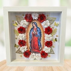 an image of the virgin mary surrounded by red and white flowers in a shadow box