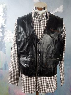 "This Swedish vintage black leather motorcycle vest has a V neckline, and closes in the front with four silvery black snap buttons. The brand name Halvarssons is embroidered on the left chest, and the handsome genuine leather waistcoat has two buttoned pockets on the front. Each lower side has an adjustable leather mini-belt. The back features a stunning embroidered motorcycle tire with the brand name running through the center of it. The vest is fully lined in a beige cotton fabric. Chest = 42 Black Biker Jacket With Snap Buttons, Sleeveless Black Biker Jacket For Fall, Biker Leather Jacket With Buttons, Sleeveless Black Biker Jacket For Biker Events, Black Moto Vest For Fall, Moto Vest For Biker Events In Fall, Moto Style Vest For Biker Events In Fall, Black Rockabilly Biker Jacket For Biker Events, Leather Vest Jacket For Fall Biker Events