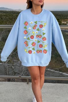 Young woman with brown hair wearing an oversized light blue crewneck sweatshirt with a print of strawberries and strawberry blossoms. She is standing on the side of a road in front of a peach and blue sunset. Long Sleeve Cotton Tops With Strawberry Print, Long Sleeve Strawberry Print Spring Tops, Long Sleeve Tops With Strawberry Print For Spring, Spring Cottagecore, Strawberry Aesthetic, Retro Cottagecore, Cottagecore Sweater, Gifts For Gardeners, Cottagecore Gifts