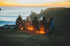 four people sitting on the edge of a cliff with candles in their hands and looking out at the ocean