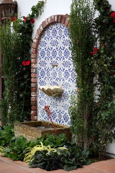 a fountain surrounded by greenery and flowers in front of a wall with an arched door