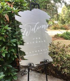 a welcome sign sitting on top of a sidewalk next to a lush green bush and shrubbery