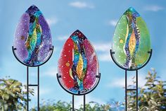 three colorful stained glass pieces on metal poles with trees in the back ground and blue sky behind them