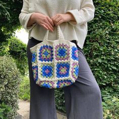 a woman holding a crocheted bag in front of her legs and wearing grey pants