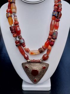 Carnelian, Agate & Trade Bead Double Strand Pendant Necklace with triangular Afghan center pendant with bezel set Carnelian stone inlays. Necklace is 18.5 inches in length.  The center pendant is 68 mm wide (at widest).  Excavation, silver, Aja and Trade Beads included.  Note: colors are a bit more subtle than they appear in the photos. Beaded Amulet Necklaces With Semi-precious Stone, Unique Carnelian Necklace With Large Pendant, Unique Red Carnelian Necklaces, Red Carnelian Amulet Necklace, Carnelian Amulet Necklace With Natural Stones, Traditional Carnelian Gemstone Necklaces, Carnelian Orange Amulet Necklace, Orange Carnelian Amulet Necklace, Red Carnelian Artisan Necklace