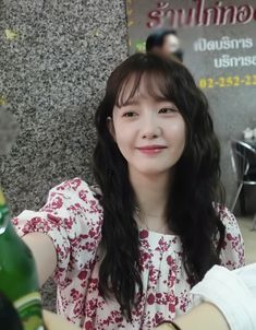 a woman with long black hair sitting next to a wall and holding a wine bottle