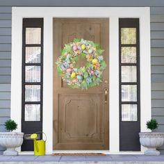 a wreath on the front door of a house with potted plants and yellow watering can