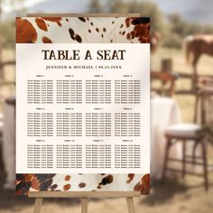 a table seating sign with brown and white spotted cow print on it in front of a farm setting