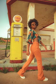 a woman in an orange pants and floral top standing next to a yellow gas pump