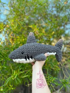 a crocheted shark toy is held up in front of some green trees and bushes