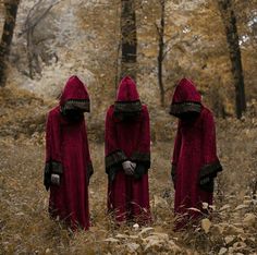 three people in red cloaks are standing in the woods with their backs to each other