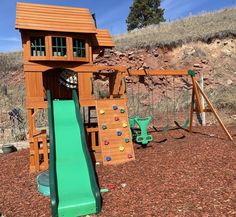 a wooden play set with a green slide and climbing frame in the background, on dirt ground