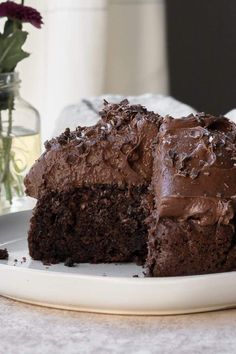 a piece of chocolate cake on a white plate with a vase of flowers in the background