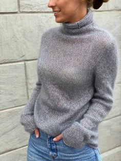 a woman standing in front of a brick wall wearing a gray turtle neck sweater and jeans