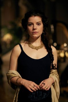 a woman with long hair wearing a black dress and gold jewelry standing in a dark room