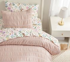 a bed with pink comforter and pillows in a room next to a white nightstand