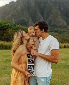 a man and woman holding a baby while standing in a field with mountains behind them