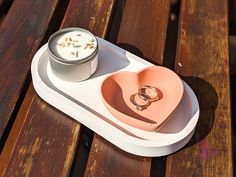 two wedding rings are placed in a heart shaped dish on a wooden table with a candle