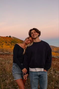 a man and woman standing next to each other in a field with trees behind them