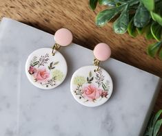 two white and pink flowered earrings on top of a marble table next to a potted plant