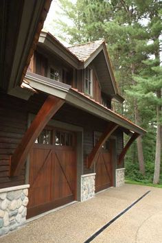 two garages with wooden doors and windows in front of a wooded area that has pine trees on the other side