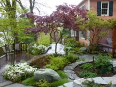 a garden with rocks and trees in front of a house next to a walkway that leads to a deck