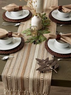 the table is set with white plates and silverware, brown napkins and leaves