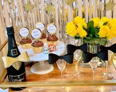 a table topped with cupcakes and wine glasses filled with yellow roses next to a bottle of champagne