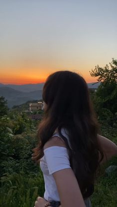 a woman standing on top of a lush green field next to a forest at sunset