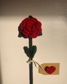 a crocheted red rose with a heart hanging on the wall next to a piece of paper