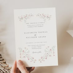 a person holding up a wedding card in front of flowers and greenery on a table