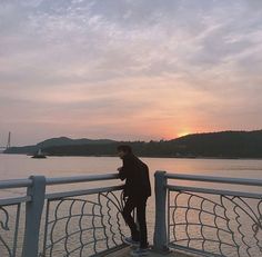 a man standing on top of a pier next to the ocean at sunset with a sailboat in the distance