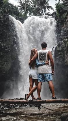 a man and woman standing on a log in front of a waterfall with water cascading