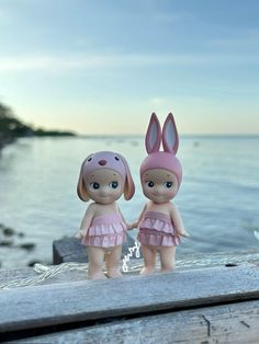 two little dolls are standing next to each other near the water, one is wearing a pink dress