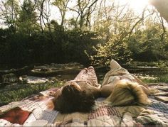 a woman laying on top of a blanket in the grass next to a river and trees